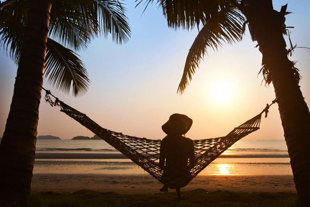silhouette of woman in hat sitting alone in hammock at sunset on the beach